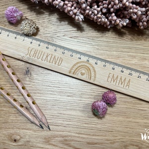 May include: A wooden ruler with a rainbow design and the words "SCHULKIND" and "EMMA" engraved on it. The ruler is 25 inches long and has markings for each inch. Two pencils with yellow polka dots are lying on a wooden surface next to the ruler.