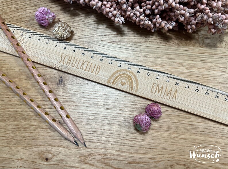May include: A wooden ruler with a rainbow design and the words "SCHULKIND" and "EMMA" engraved on it. The ruler is 25 inches long and has markings for each inch. Two pencils with yellow polka dots are lying on a wooden surface next to the ruler.