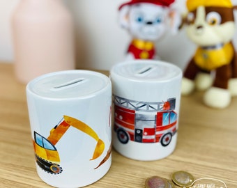 May include: Two white ceramic piggy banks with different designs. One has a red fire truck and the other has a yellow excavator. Both piggy banks have a slot on top for coins.