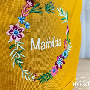 K&ouml;nnte beinhalten: Eine gelbe Stofftasche mit einer gestickten Blumenkrone um den Namen "Mathilda".