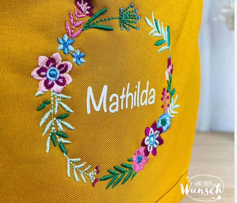 K&ouml;nnte beinhalten: Eine gelbe Stofftasche mit einer gestickten Blumenkrone um den Namen "Mathilda".