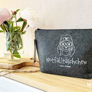 May include: A gray felt pouch with a zipper closure and a white graphic of a rabbit wearing glasses and a hat. The text "Notfallt&auml;schchen VON JONAS" is printed below the graphic. A vase of pink peonies sits to the left.