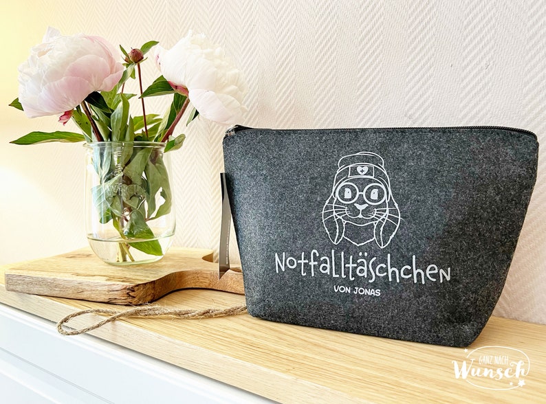 May include: A gray felt pouch with a zipper closure and a white graphic of a rabbit wearing glasses and a hat. The text "Notfallt&auml;schchen VON JONAS" is printed below the graphic. A vase of pink peonies sits to the left.
