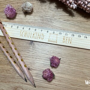 May include: A wooden ruler with a cute bear illustration and the text "SCHULKIND BEN" engraved on it. The ruler is 16 inches long and has markings for each inch. Two pencils with gold polka dots are lying on a wooden surface next to the ruler.
