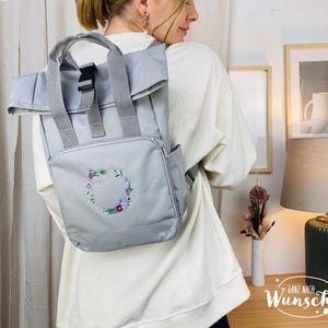 K&ouml;nnte beinhalten: Ein grauer Rucksack mit Rollverschluss und einer gestickten Blumenkrone auf der Vorderseite. Der Rucksack hat eine schwarze Schnalle und verstellbare Tr&auml;ger. Der Text "Ganz nach Wunsch" ist in der unteren rechten Ecke des Bildes sichtbar.