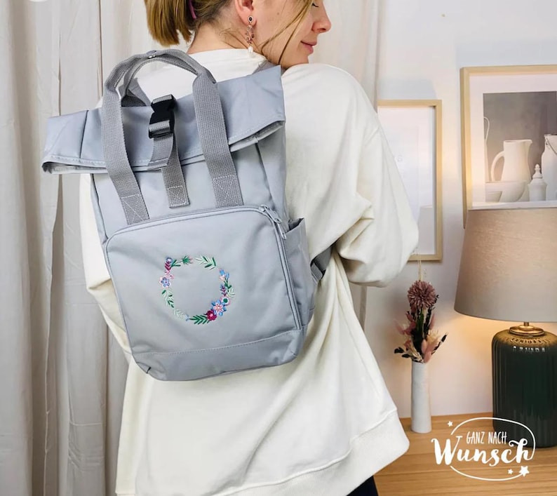 K&ouml;nnte beinhalten: Ein grauer Rucksack mit Rollverschluss und einer gestickten Blumenkrone auf der Vorderseite. Der Rucksack hat eine schwarze Schnalle und verstellbare Tr&auml;ger. Der Text "Ganz nach Wunsch" ist in der unteren rechten Ecke des Bildes sichtbar.