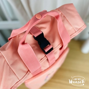 K&ouml;nnte beinhalten: Ein rosa Rucksack mit einer schwarzen Schnalle und einem rosa Gurt. Der Gurt hat einen rosa Knopf oben.