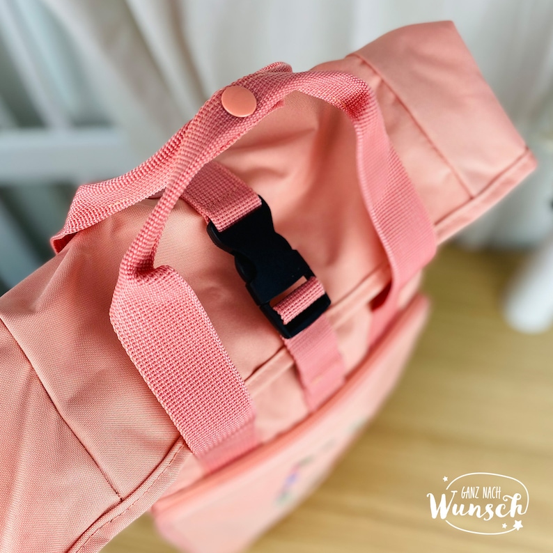 K&ouml;nnte beinhalten: Ein rosa Rucksack mit einer schwarzen Schnalle und einem rosa Gurt. Der Gurt hat einen rosa Knopf oben.