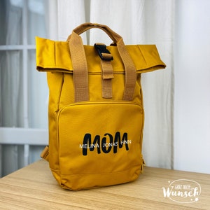Könnte beinhalten: Ein senfgelber Rucksack mit schwarzen Trägern und einer schwarzen Schnalle. Der Rucksack hat ein großes Fach auf der Vorderseite mit dem Wort "MOM" in schwarzen Buchstaben und den Namen "MELINA JONAS FINN" darunter in kleineren weißen Buchstaben.