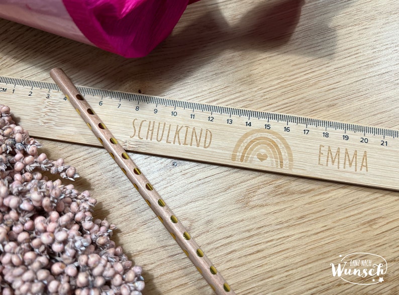 May include: A wooden ruler with a rainbow design and the words "SCHULKIND" and "EMMA" engraved on it. The ruler is 21 inches long and has markings for each inch. A yellow pencil with black dots is lying next to the ruler.