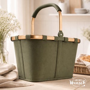 Pu&ograve; includere: Un cestino verde oliva con una finitura liscia e opaca e una struttura in metallo color oro. Il cestino presenta un manico ad arco singolo con un'impugnatura abbinata in verde oliva e una base rettangolare. Il testo "Ganz nach Wunsch" &egrave; stampato in bianco.