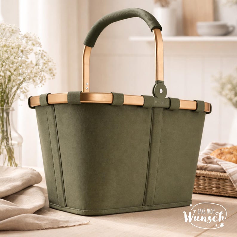 Pu&ograve; includere: Un cestino verde oliva con una finitura liscia e opaca e una struttura in metallo color oro. Il cestino presenta un manico ad arco singolo con un'impugnatura abbinata in verde oliva e una base rettangolare. Il testo "Ganz nach Wunsch" &egrave; stampato in bianco.