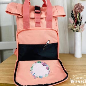 K&ouml;nnte beinhalten: Ein pfirsichfarbener Rucksack mit einem Klappdeckel und einer schwarzen Rei&szlig;verschlusstasche. Der Rucksack hat ein florales Stickmuster auf der vorderen Klappe und den Text "Ganz nach Wunsch" unten.