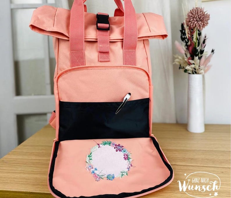 K&ouml;nnte beinhalten: Ein pfirsichfarbener Rucksack mit einem Klappdeckel und einer schwarzen Rei&szlig;verschlusstasche. Der Rucksack hat ein florales Stickmuster auf der vorderen Klappe und den Text "Ganz nach Wunsch" unten.