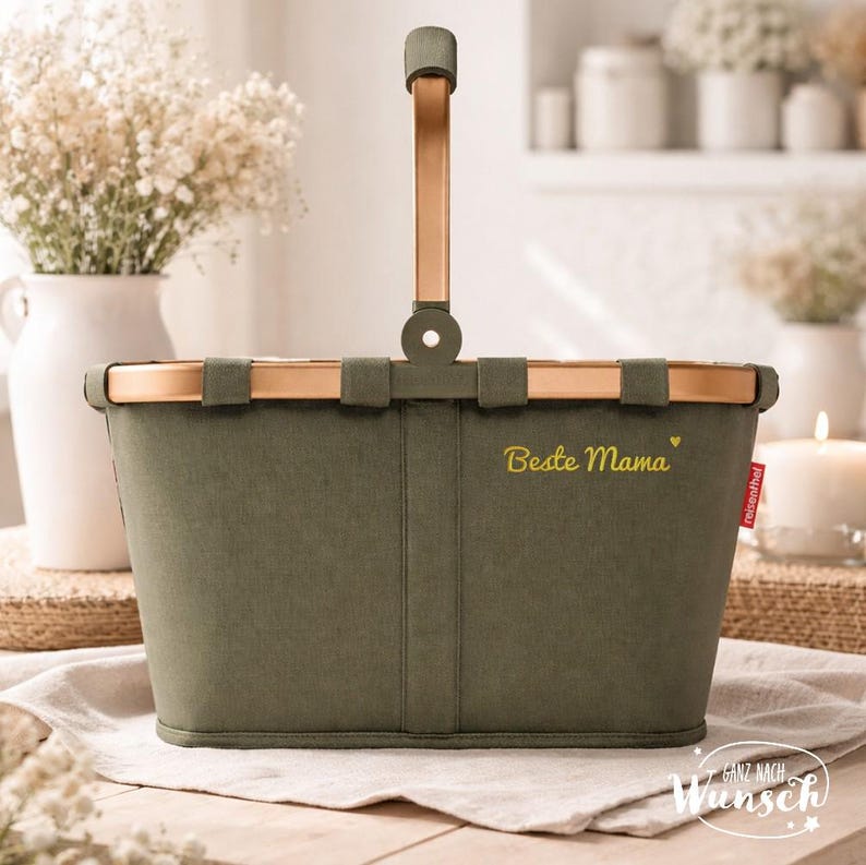 Op de afbeelding: Een olijfgroene stoffen mand met een ros&eacute;gouden metalen frame en een enkele rechtopstaande handgreep. De voorzijde is voorzien van de geborduurde gele tekst "Beste Mama" met een klein hartje en een rood "reisenthel"-label aan de zijkant.