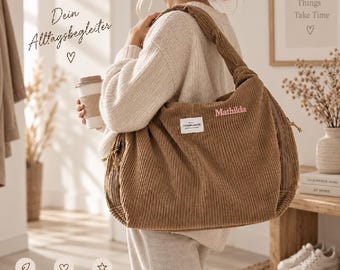 May include: A brown corduroy shoulder bag featuring a knotted strap, a white rectangular label, and the name Mathilda embroidered in pink thread. The bag has a textured ribbed surface, side zippers with pull cords, and a spacious main compartment.