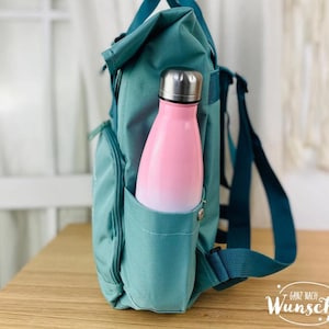 K&ouml;nnte beinhalten: Tealblauer Rucksack mit einer Seitentasche, in der eine rosa-wei&szlig;e Wasserflasche steckt. Der Rucksack hat einen tealblauen Gurt und der Text "Ganz nach Wunsch" ist in der rechten unteren Ecke des Bildes zu sehen.
