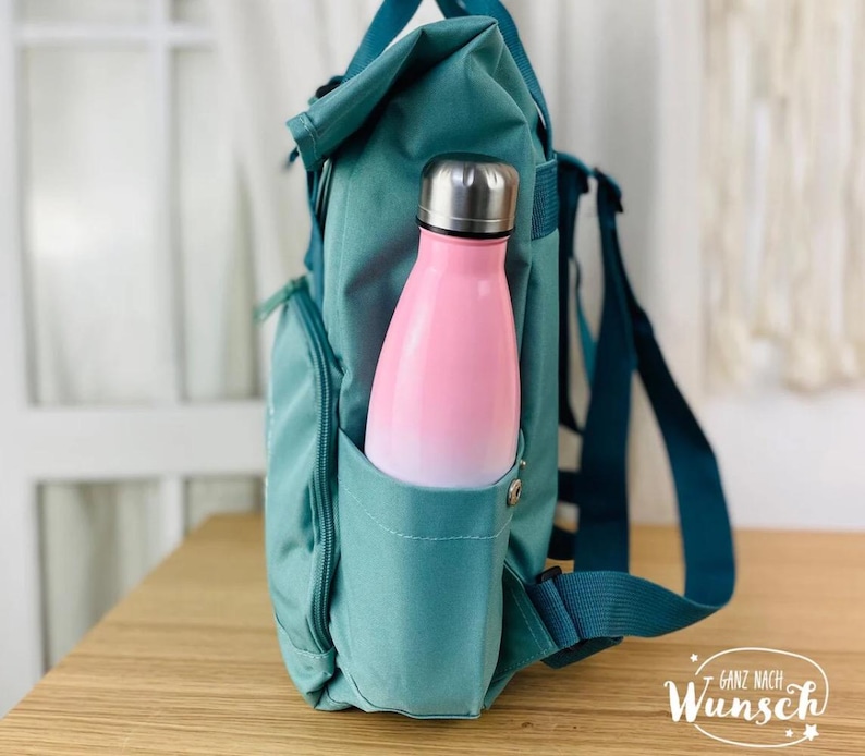 K&ouml;nnte beinhalten: Tealblauer Rucksack mit einer Seitentasche, in der eine rosa-wei&szlig;e Wasserflasche steckt. Der Rucksack hat einen tealblauen Gurt und der Text "Ganz nach Wunsch" ist in der rechten unteren Ecke des Bildes zu sehen.