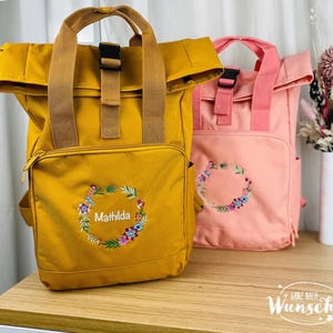 Könnte beinhalten: Zwei personalisierte Rucksäcke, einer in Senfgelb und einer in Hellrosa, mit Blumenstickerei und dem Namen "Mathilda" auf dem gelben Rucksack. Die Rucksäcke haben einen Rollverschluss und verstellbare Träger.