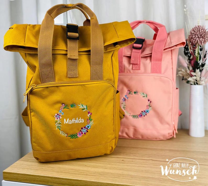 K&ouml;nnte beinhalten: Zwei personalisierte Rucks&auml;cke, einer in Senfgelb und einer in Hellrosa, mit Blumenstickerei und dem Namen "Mathilda" auf dem gelben Rucksack. Die Rucks&auml;cke haben einen Rollverschluss und verstellbare Tr&auml;ger.