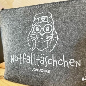 May include: Gray felt zippered pouch with a white illustration of a bunny wearing glasses and a nurse's cap. The text "Notfallt&auml;schchen VON JONAS" is printed below the bunny illustration.