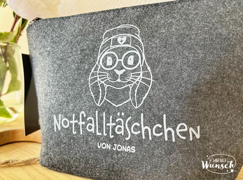 May include: Gray felt zippered pouch with a white illustration of a bunny wearing glasses and a nurse's cap. The text "Notfallt&auml;schchen VON JONAS" is printed below the bunny illustration.
