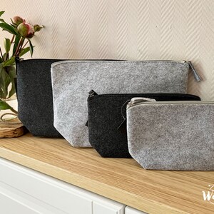 May include: Four felt pouches in varying sizes and shades of gray and black are arranged on a wooden surface. Each pouch has a zipper closure. A glass vase with flowers sits to the left of the pouches.