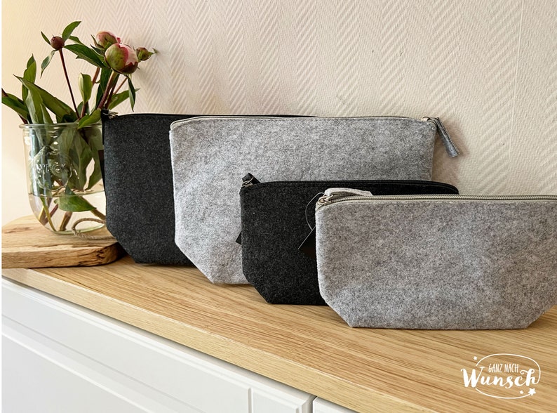 May include: Four felt pouches in varying sizes and shades of gray and black are arranged on a wooden surface. Each pouch has a zipper closure. A glass vase with flowers sits to the left of the pouches.
