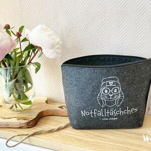 May include: A gray felt zippered pouch with a white cartoon rabbit design wearing a hat and glasses. The text "Notfallt&auml;schchen von Jonas" is printed below the rabbit. A glass vase with pink and white peonies sits to the left.