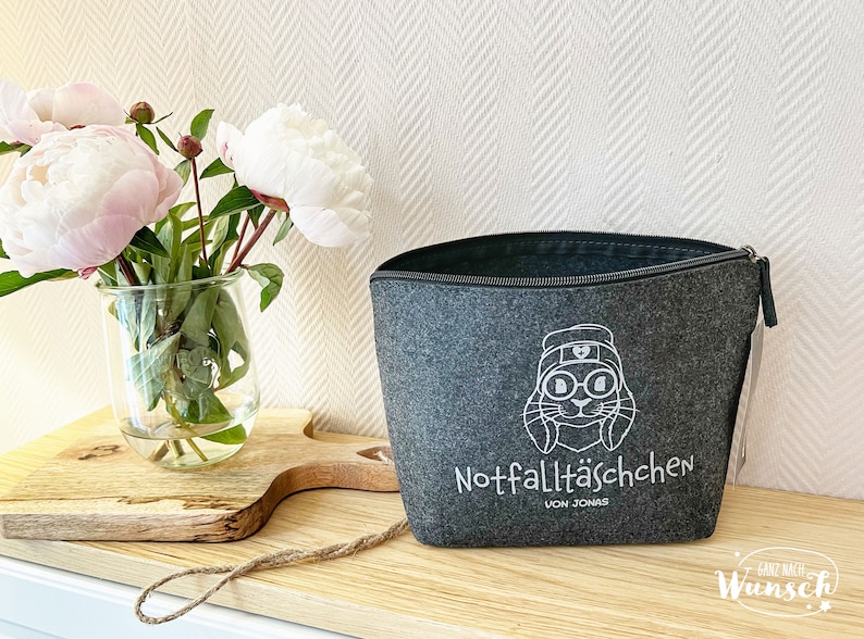 May include: A gray felt zippered pouch with a white cartoon rabbit design wearing a hat and glasses. The text "Notfallt&auml;schchen von Jonas" is printed below the rabbit. A glass vase with pink and white peonies sits to the left.