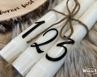 May include: Three white candles with black numbers 1, 2, and 3 printed on them. The candles are tied together with twine.