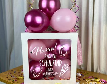 May include: A white box with a clear window filled with pink and purple balloons. The box has a white sticker with the text "Hurra! Endlich Schulkind Imke 10. August 2024".