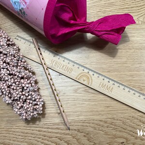 May include: A wooden ruler with a rainbow design and the words "SCHULKIND" and "EMMA" engraved on it. The ruler is 30 cm long and has a pencil next to it.