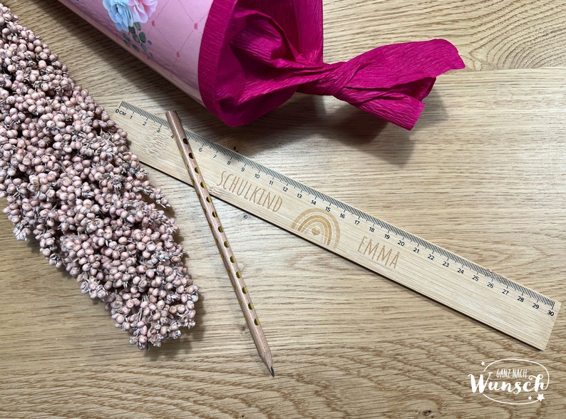 May include: A wooden ruler with a rainbow design and the words "SCHULKIND" and "EMMA" engraved on it. The ruler is 30 cm long and has a pencil next to it.