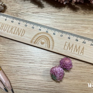 May include: A wooden ruler with a rainbow design and the word "SCHULKIND" and the name "EMMA" engraved on it. The ruler is 23 inches long and has markings for each inch.