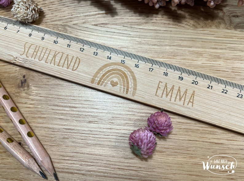 May include: A wooden ruler with a rainbow design and the word "SCHULKIND" and the name "EMMA" engraved on it. The ruler is 23 inches long and has markings for each inch.