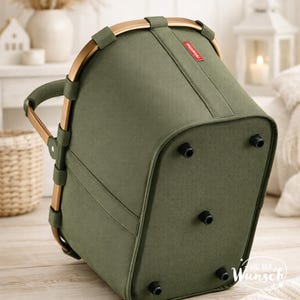 Pu&ograve; includere: Un cestino per la spesa verde oliva con manico e telaio in metallo color oro. Il cestino presenta una base rettangolare con quattro piedini in plastica nera sul fondo e una piccola etichetta rossa con il testo bianco reisenthel sul lato.