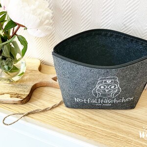May include: A gray felt zippered pouch with a white cartoon rabbit graphic and the text "Notfallt&auml;schchen". The pouch is open, revealing a dark interior. A vase of flowers sits to the left.