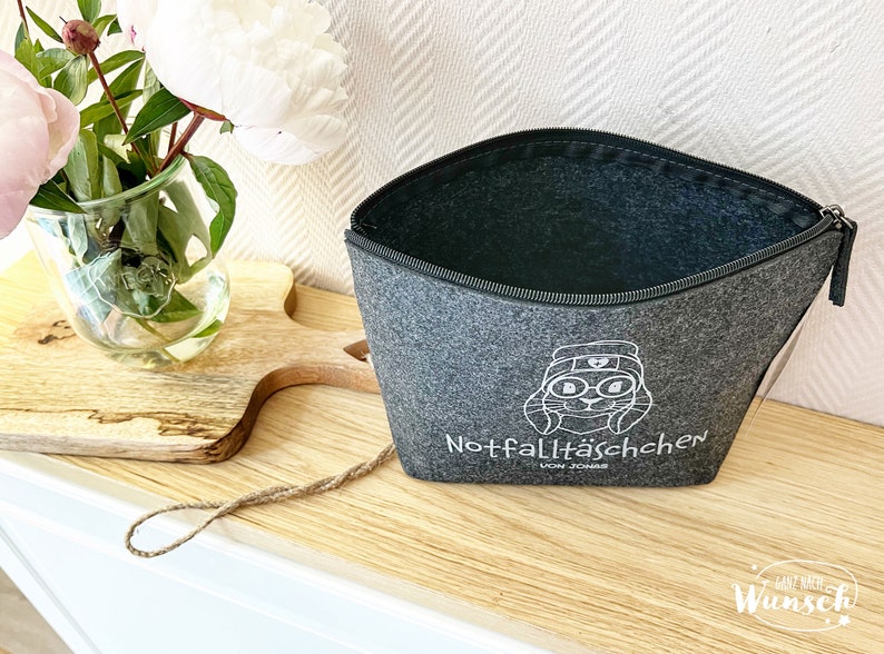 May include: A gray felt zippered pouch with a white cartoon rabbit graphic and the text "Notfallt&auml;schchen". The pouch is open, revealing a dark interior. A vase of flowers sits to the left.