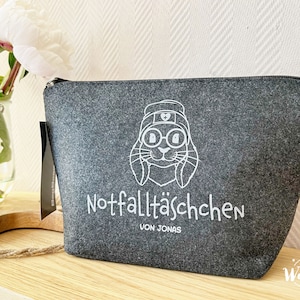 May include: Gray felt zippered pouch with a white graphic of a bunny wearing glasses and a nurse's hat. The text "Notfallt&auml;schchen" and "Von Jonas" are printed below the bunny. A white peony flower is in the background.