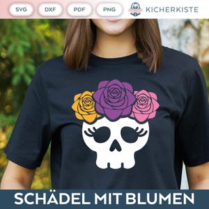 Op de afbeelding: Een zwart T-shirt met een witte schedel die een bloemenkrans draagt. De bloemen zijn geel, paars en roze. De tekst "SCHÄDEL MIT BLUMEN" is onder de schedel gedrukt.