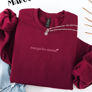 May include: Maroon crewneck sweatshirt with the text "margarita mama" embroidered in a light color. A silver necklace with a pendant is draped on the sweatshirt. The word "MAROON" is printed in black at the top.