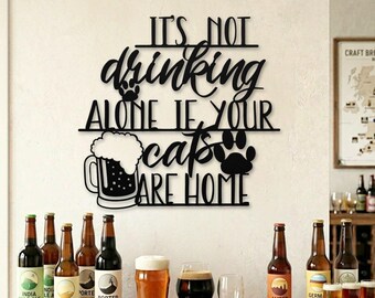 May include: A black metal sign with the text "IT'S NOT drinking ALONE IF YOUR cats ARE HOME" above a beer mug and paw prints. Various beer bottles and glasses are arranged on a wooden surface, suggesting a home bar or pub setting.