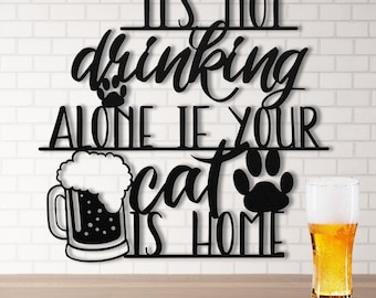 May include: Black metal wall art with the phrase "IT'S NOT drinking ALONE IF YOUR cat IS HOME". The design includes a beer mug, cat paw prints, and a glass of beer. The background is a white brick wall.