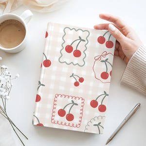 May include: A notebook with a cherry pattern. The cover features a light beige gingham background with red cherries in various decorative frames. A white coffee mug and a pen are also visible.