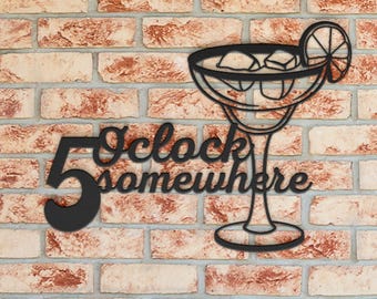May include: Black metal wall art featuring a margarita glass with ice cubes and a lime slice. The phrase "5 O'clock somewhere" is displayed in a decorative font. The art is mounted on a brick wall.