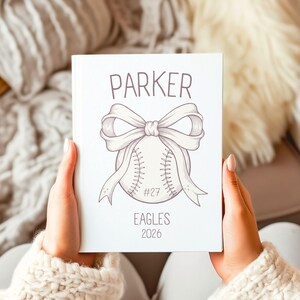 May include: A white notebook featuring a baseball design with a bow and the name "PARKER". Below the baseball are the words "EAGLES" and the year "2026". The notebook is held by a person wearing a cream-colored sweater.