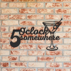 May include: Black metal wall art featuring a martini glass and the phrase "5 O'clock somewhere" in a decorative font. The artwork includes an olive on a toothpick. The background is a brick wall.