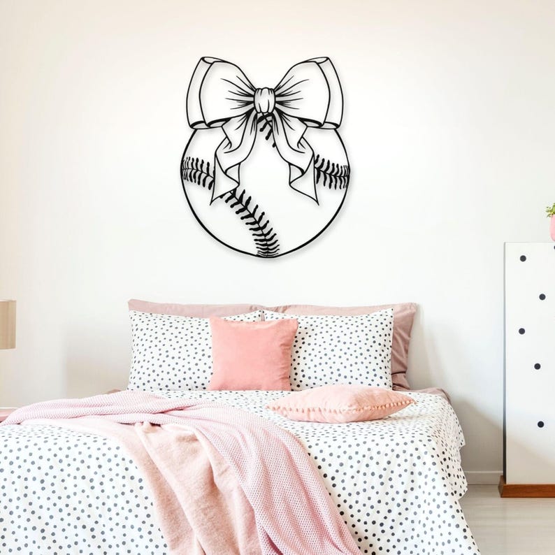 May include: A black and white illustration of a baseball with a large bow on top. The illustration is on a white wall above a bed with pink and white polka dot bedding.