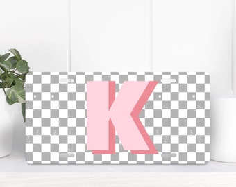Puede incluir: Una placa de matrícula rectangular con una letra K de color rosa claro centrada sobre un fondo de cuadros blancos y grises. La letra K tiene un efecto de sombra rosa más oscuro y el patrón de cuadros tiene una textura desgastada.